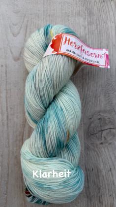 Vier handgefärbte Garnstränge der Marke Herzfasern liegen nebeneinander auf hellem Untergrund. Der Strang „Klarheit“ zeigt eine ruhige, helle Farbgebung mit sanften, klaren Nuancen. Die Farben wirken offen, leicht und ausgewogen.